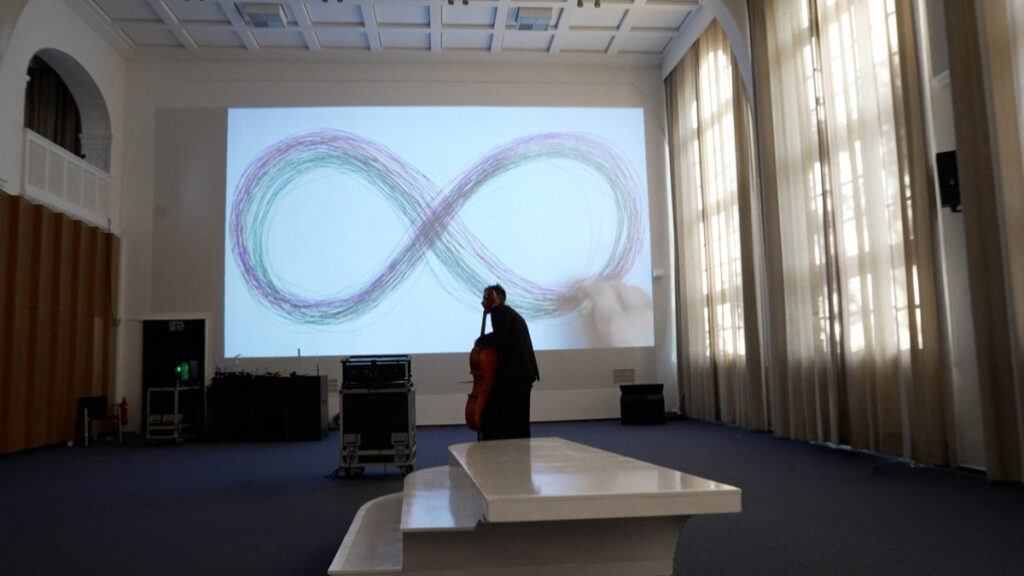 Whilst a cellist is standing on an interactive sculptural interface, a video of a hand drawing an horizontal eight is projected on almost an entire rastered wall in the auditorium of LI Hamburg.