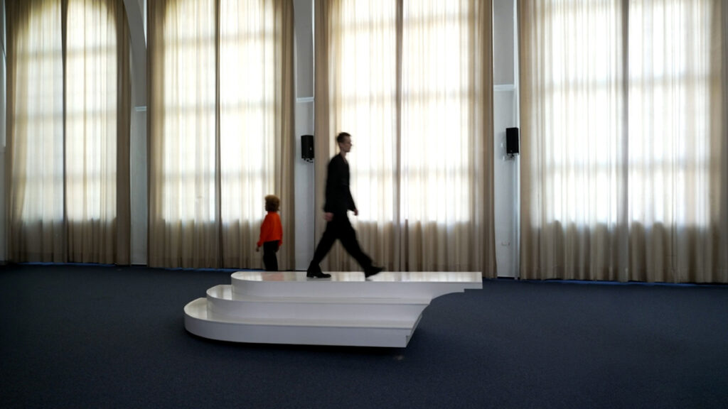 3 performers keep crossing an interactive sea-saw sculptural interface in a choreographed manner, with different speeds and attitudes, sometimey apruptly changing directions. The sculpture has a rectangular, platform like shape, with three steps at the sides.