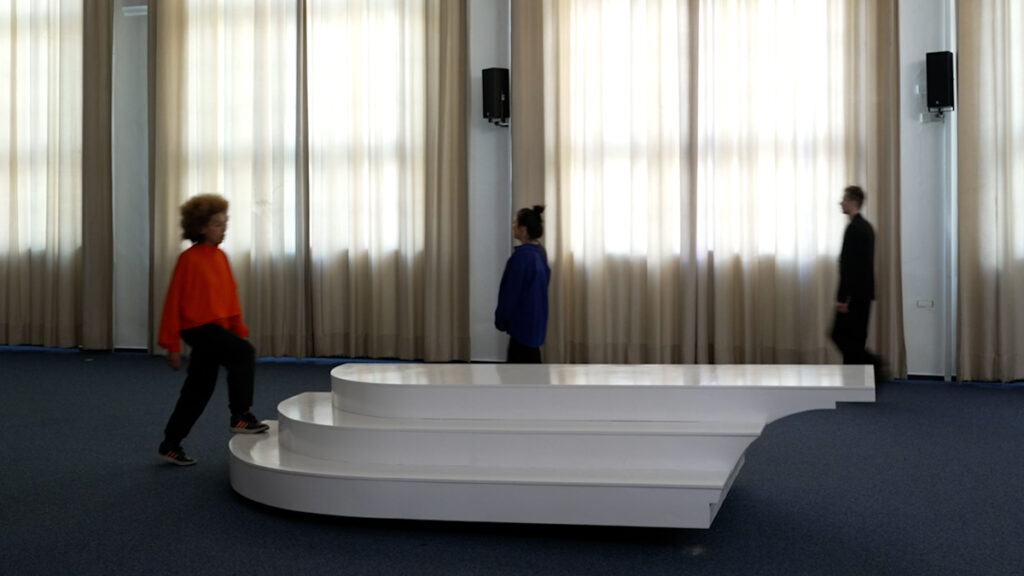 3 performers keep crossing an interactive sea-saw sculptural interface in a choreographed manner, with different speeds and attitudes, sometimey apruptly changing directions. The sculpture has a rectangular, platform like shape, with three steps at the sides.