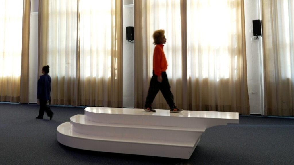 3 performers keep crossing an interactive sea-saw sculptural interface in a choreographed manner, with different speeds and attitudes, sometimey apruptly changing directions. The sculpture has a rectangular, platform like shape, with three steps at the sides.