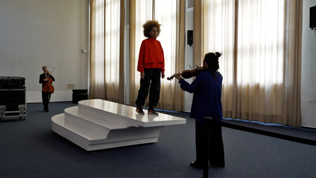 2 string instrument performers play on two ends of a white sculptural sea saw platform, with a conductor on top facing one performer.
