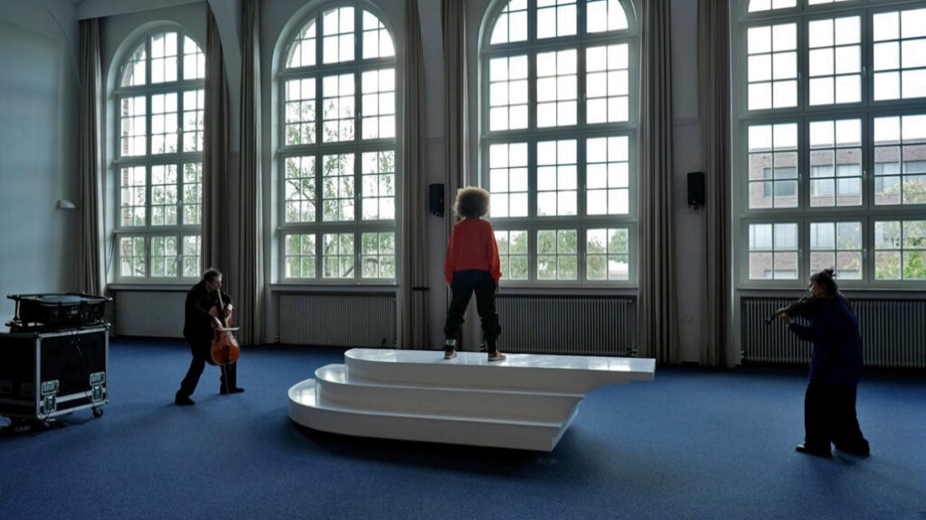 2 string instrument performers play on two ends of a white sculptural sea saw platform, with a conductor on top facing one performer.