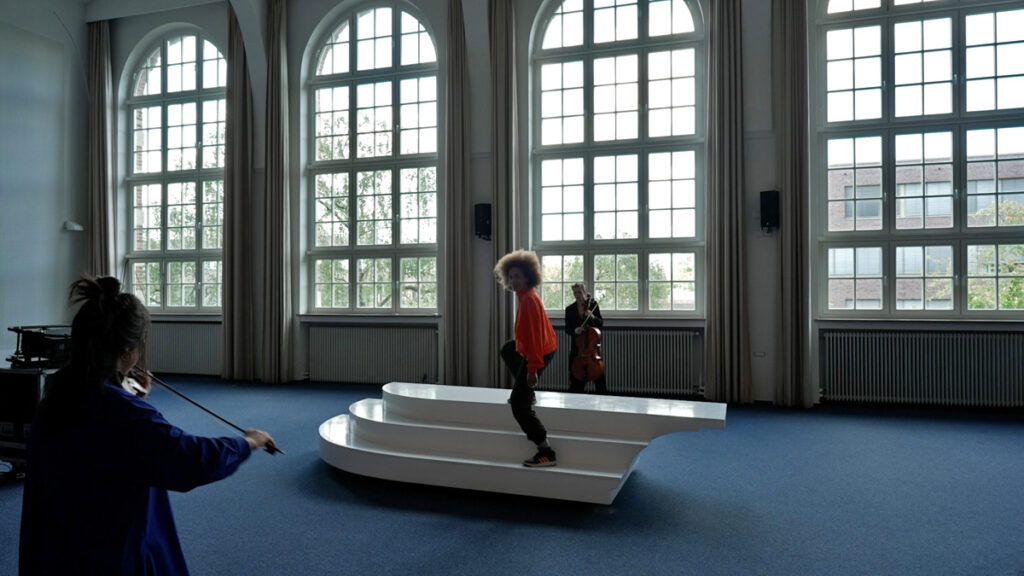 2 string instrument performers play on two ends of a white sculptural sea saw platform, with a conductor on top facing one performer.