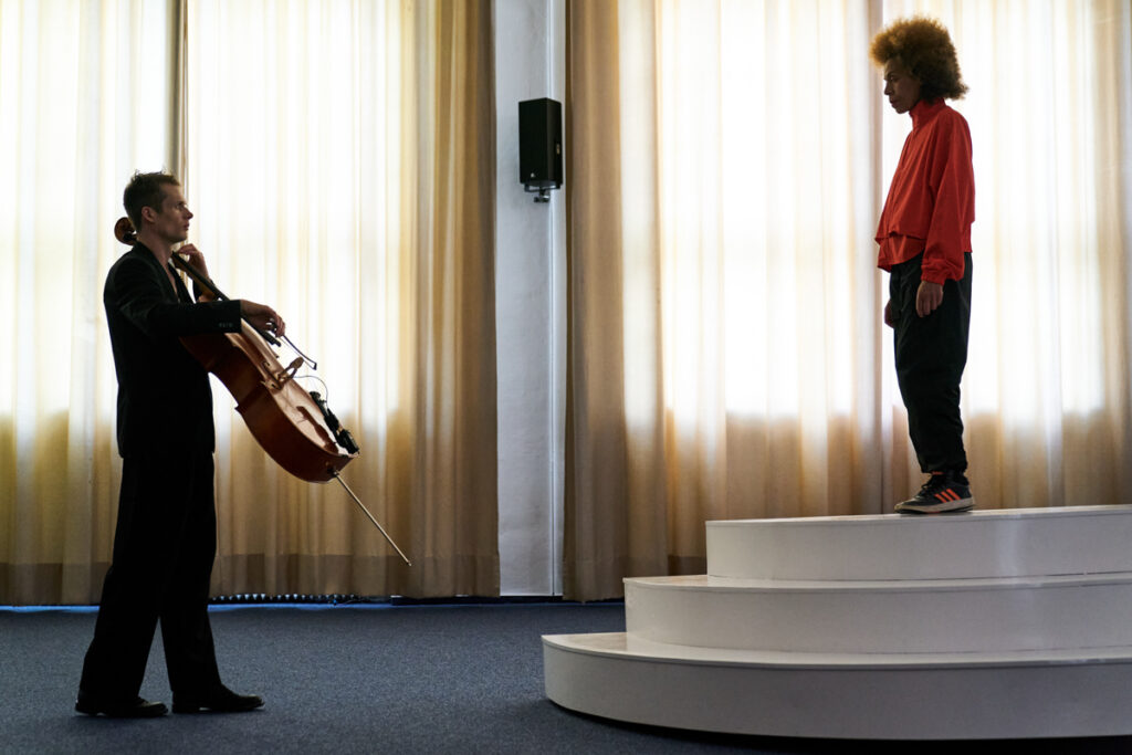 2 string instrument (cello & violin) performers play on two ends of a white sculptural sea-saw platform, with a conductor on top facing one performer.