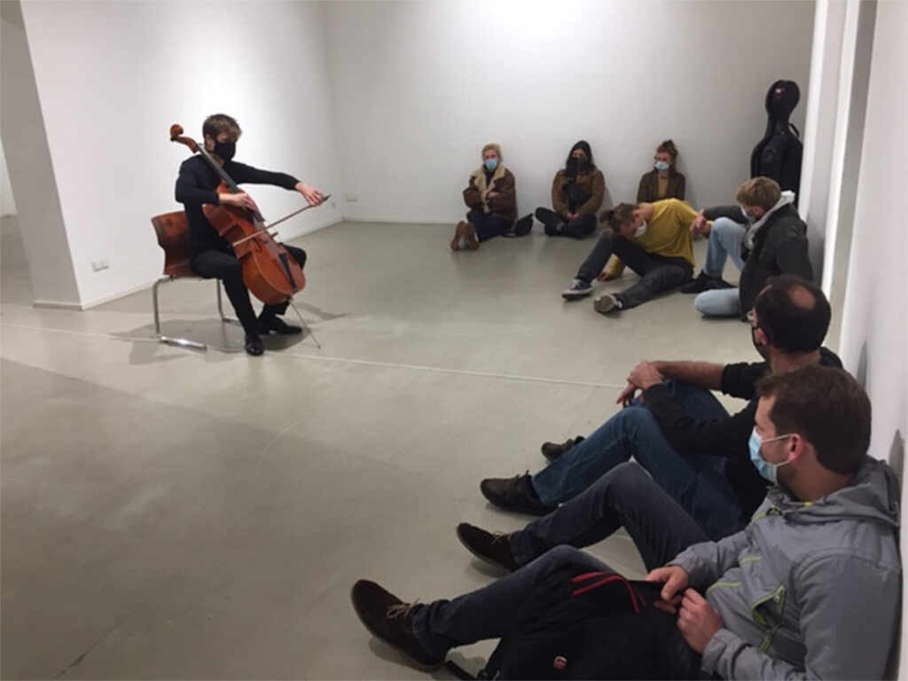 Cellist playing in empty gallery space to audience seated on the floor together with a dancer and performer interacting.