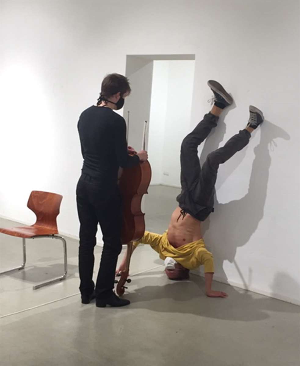 Cellist holding Cello upside down, facing dancer leaning at the wall in upside down body position. 