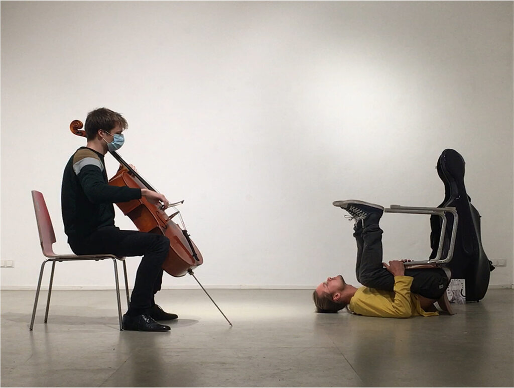 Cellist (Christopher Loy) playing seated with dancer (Nils Löfke) lying on the floor holding a chair upside down with his legs.