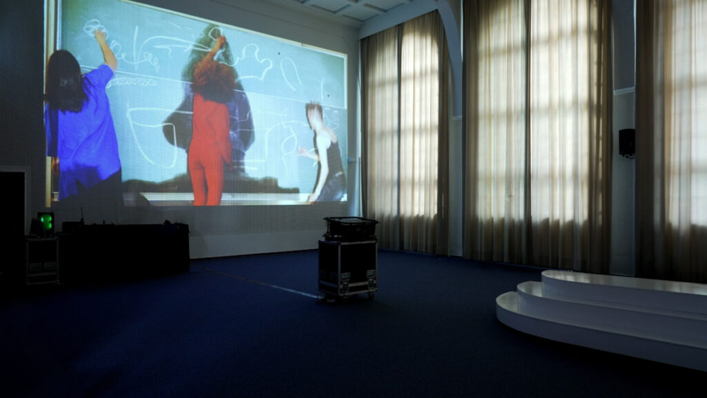 A video showing 3 performers painting over a video projected on a blackboard is projected on a wall with a raster structure in the Auditorium of LI Hamburg.