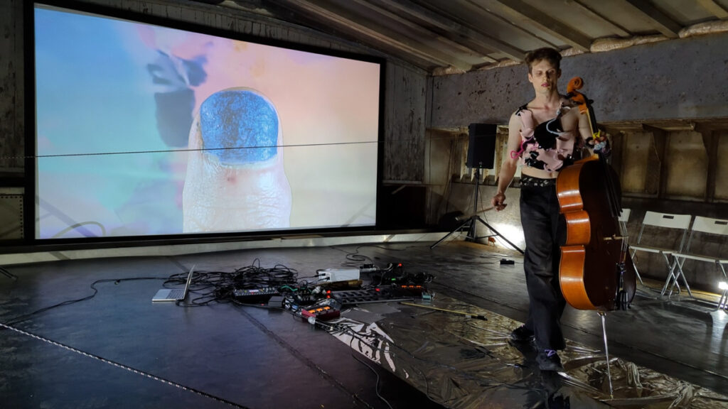 A performer with a cello is standing on a iridescent foil, whilst in the background a video with a thumb in the size of a human body is projected.