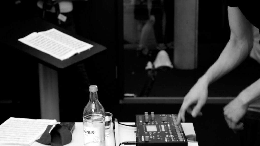 An artist is making inputs on an Octatrack Sampler interface, in the background there is a speaker's desk a little similar to a music stand with sheet music on it.