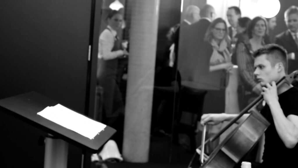 A cellist practicing in a room with a glass wall, behind which there's an opening reception happening, with people talking, drinking and eating.
