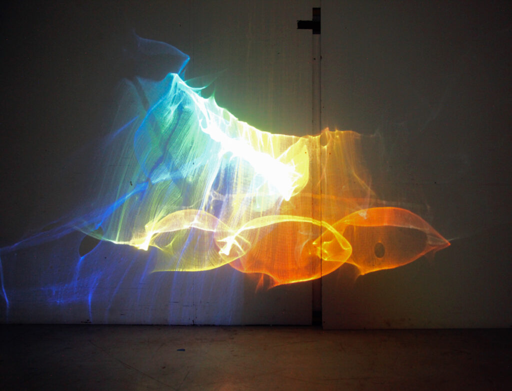 Spectral color reflection on the wall facing a video projection on transparent foil of animated pictures of light broken by glass prisms as part of a narrative room installation. 