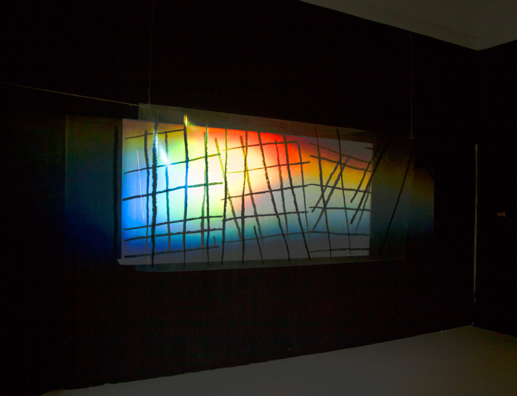 A largescale print of enlarged vertical pencil lines on transparent film is hanging in front of a large scale print on paper with vertical lines. Together they form a raster on which a video with spectral colors is projected.