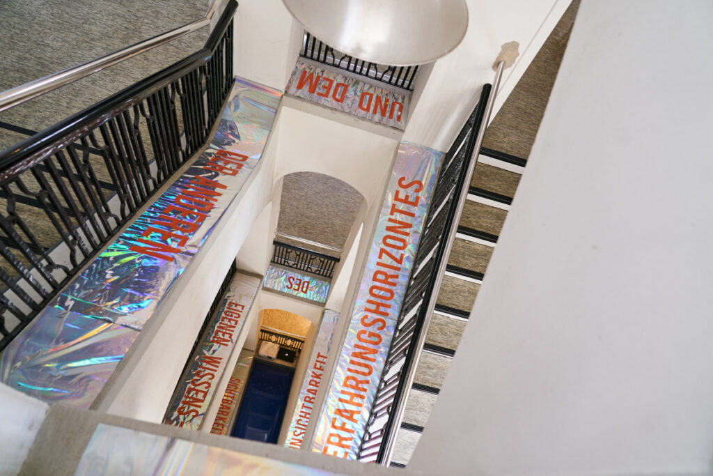 Installation of orange letters on iriddescent foil in historic Fritz-Schumacher-building staircase over three floors: Visibility & Invisibility of ones own Horizon of Knowledge and that of the other Subjects.