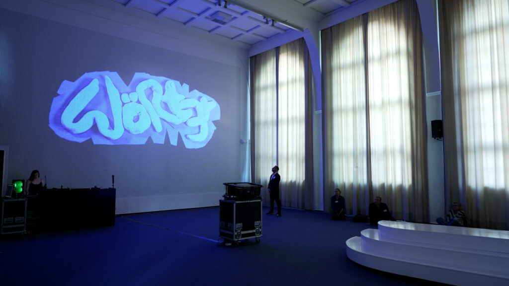 Sculptural letters in blue, saying "Wört3r" (engl. "Words") are projected on a wall with a raster structure in the auditorium of LI Hamburg.
