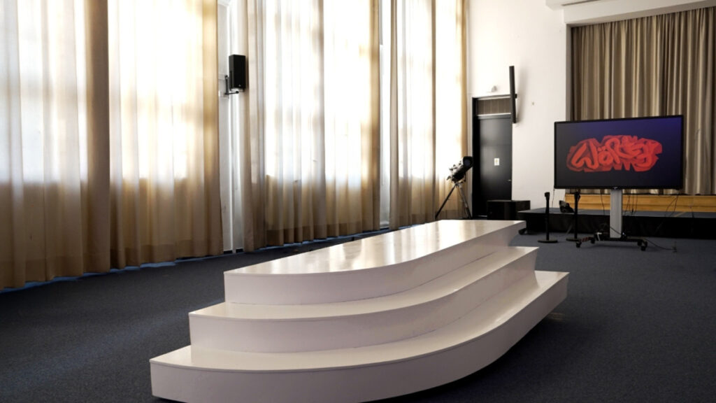 A mobile C-touch screen showing sculptural letters in red, saying "Wört3r" (engl. "Words") is standing behind a three steps, platform like seasaw sculpture.