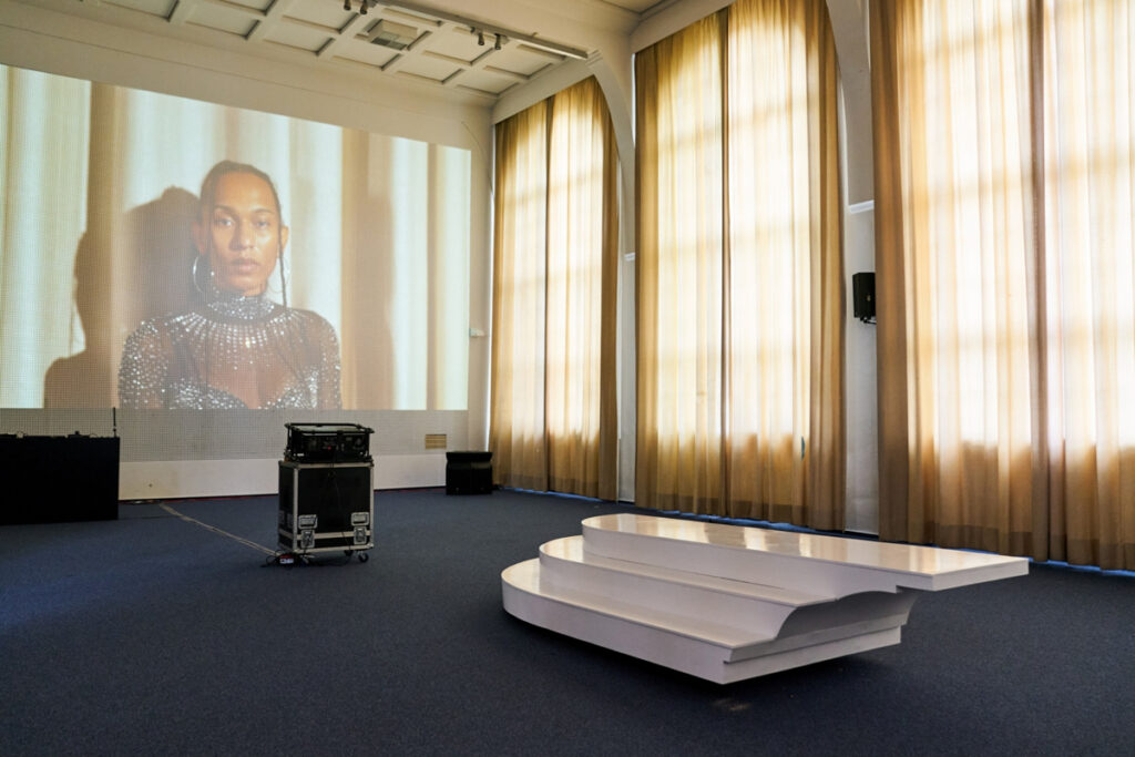 A side stepped platform is standing in the middle, in the background a close up of a female dancer is projected on a rastered wall of an empty auditorium.