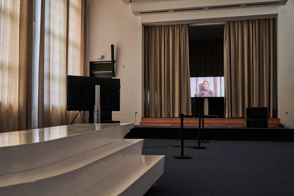 C-Touch screen on the middle of the stage of cleared LI Hamburg auditorium with curtains just opened enough to reveal the screen. The screen shows image of performer Black Pearl de Almeida Lima in close-up shot looking into the camera, filmed in front of the curtains of the very stage the screen is standing on.