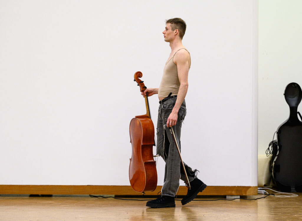 Christopher Loy, gehend mit Cello in der Hand in einem Ausstellungsraum im Museum Abteiberg