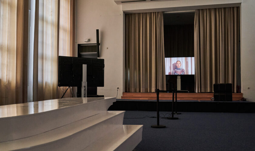 C-Touch screen on the middle of the stage of cleared LI Hamburg auditorium with curtains just opened enough to reveal the screen. The screen shows image of performer Black Pearl de Almeida Lima in close-up shot looking into the camera, filmed in front of the curtains of the very stage the screen is standing on.