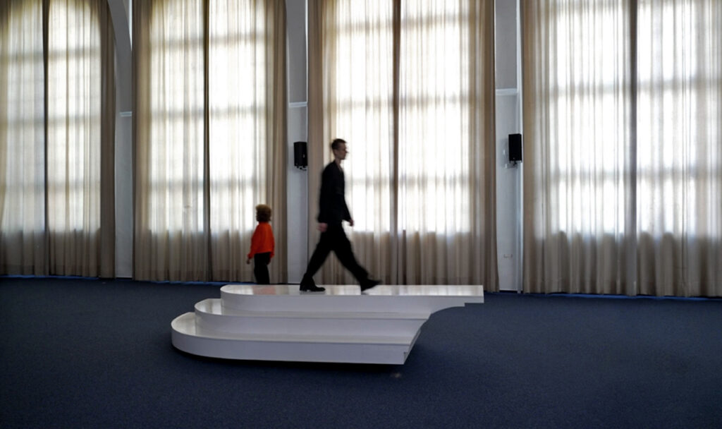 Three performers repeatedly crossing an interactive seesaw-like sculptural with staircase elements platform during choreographed movement sequence.