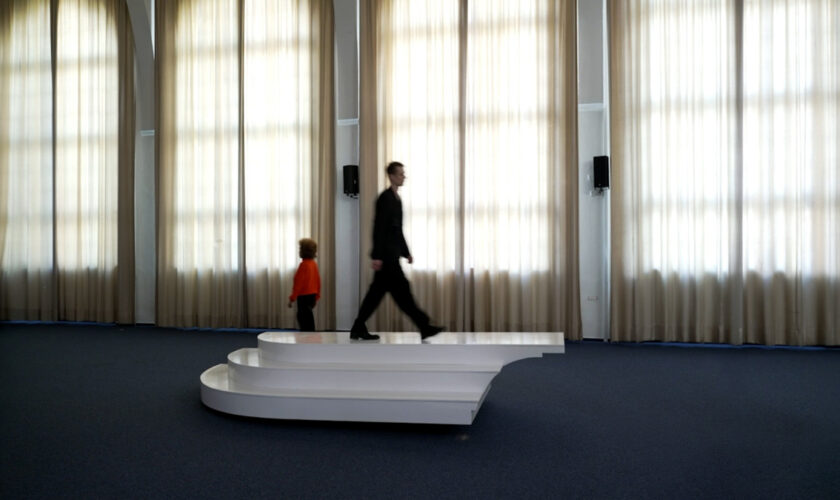 Three performers repeatedly crossing an interactive seesaw-like sculptural with staircase elements platform during choreographed movement sequence.