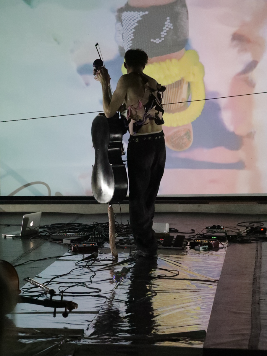 Performer playing the cello inverted with the strings facing him and one hand changing pitch by turning a peg and the other plucking a string. A video with an index finger in the size of a human body is projected in the background.