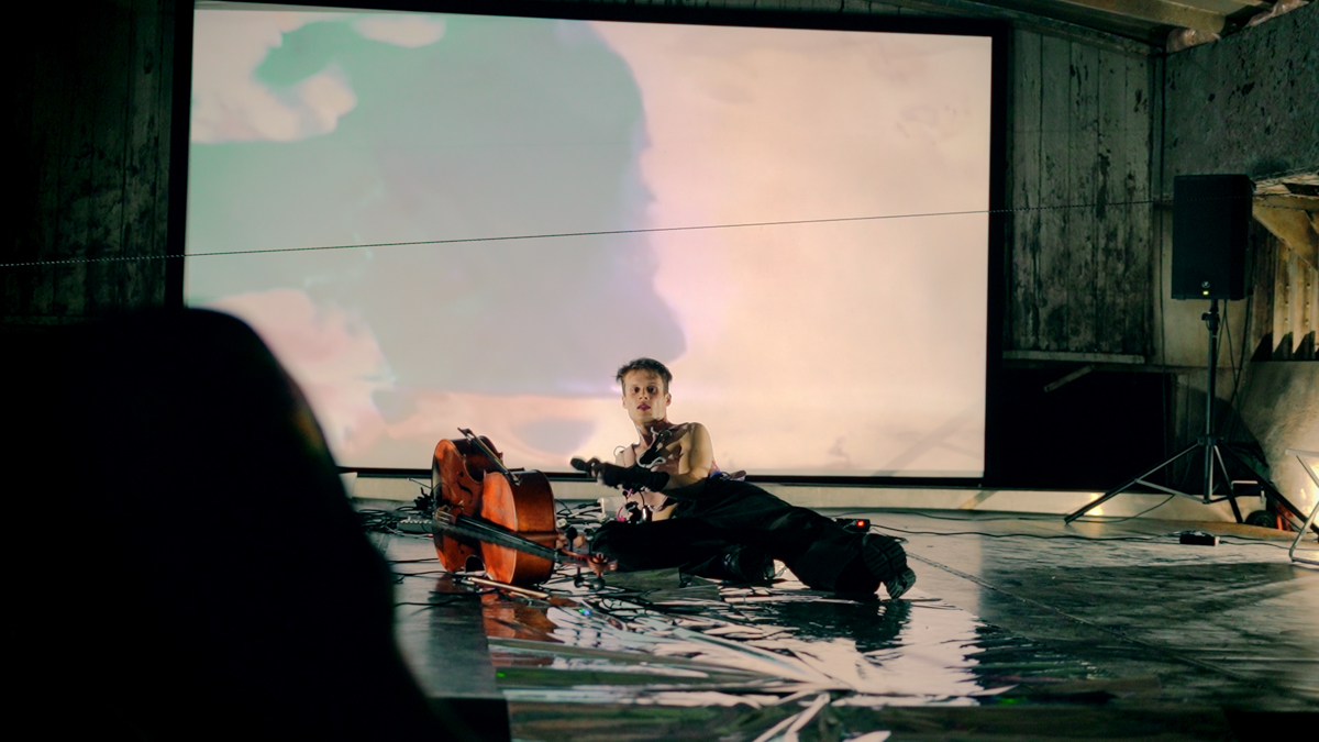 A performer is lying on an iridescent foil carpet with a cello next to him and an abstract video projection with color gradient in the background.