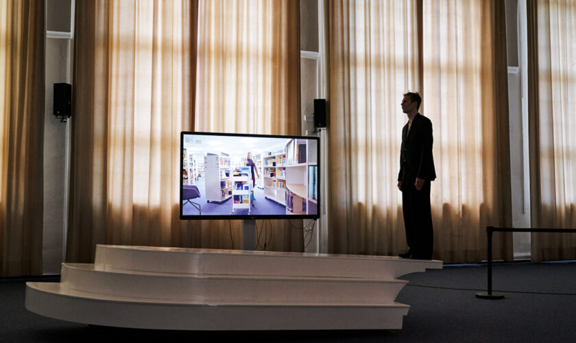 A performer is standing on the edge of an interactive sculpture in the middle of the space. In the background, a dancer, Black Pearl de Almeida Lima, artistically rearranging rolling shelfs in the LI Hamburg library in a sculptural, installative manner during night, is viewd on a screen in front of closed curtains.