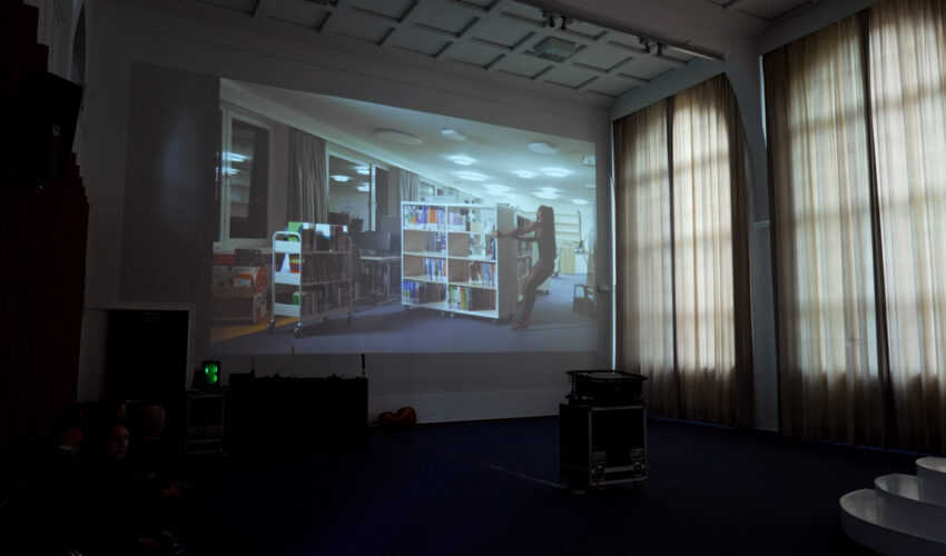 Projection on a rastered auditorium wall showing a female dancer moving rolling library shelves at night.