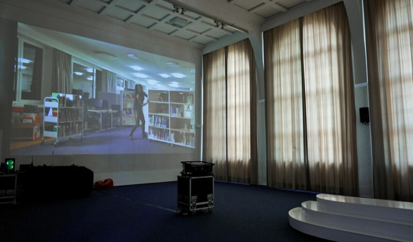 Projection on a rastered auditorium wall showing a female dancer moving rolling library shelves at night.