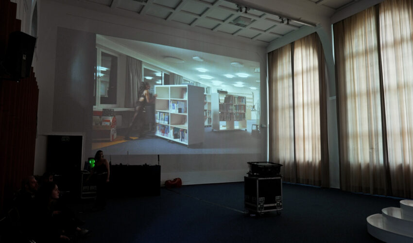 Projection on a rastered auditorium wall showing a female dancer moving rolling library shelves at night.